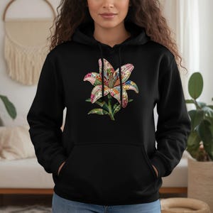 Puede incluir: Sudadera con capucha negra con un diseño floral colorido. La flor presenta un patrón detallado con varios colores, incluyendo rosa, naranja y azul. La sudadera tiene un bolsillo de canguro y una capucha con cordón.
