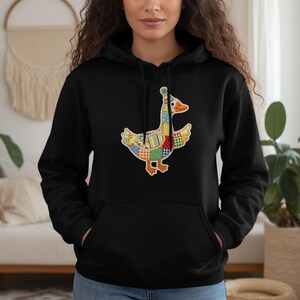 Op de afbeelding: Zwarte hoodie met een kleurrijk patchwork ganzenontwerp. De gans heeft een gele snavel, oranje poten en een hoed. Het ontwerp bevat verschillende patronen en kleuren, waaronder rood, blauw, geel en groen. De hoodie heeft een capuchon met trekkoord en een voorzak.