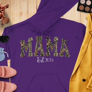 May include: A purple hoodie featuring the word "MAMA" in a camouflage pattern, with "Est. 2025" in white script underneath. The hoodie has a front pocket and a drawstring hood. Sunglasses, makeup brushes, and a yellow blazer are also visible.