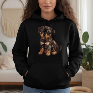 May include: Black hooded sweatshirt featuring a graphic of a Rottweiler puppy with a patchwork design. The puppy has brown, black, and tan fur and is wearing a patchwork scarf. The sweatshirt has a kangaroo pocket and a drawstring hood.