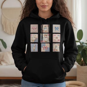 May include: Black hoodie featuring nine square patches with vintage-style illustrations. The patches showcase images of geese, bows, and floral patterns in pastel colors. The hoodie has a drawstring hood and a front pocket.