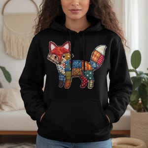 May include: Black hoodie with a colourful patchwork fox design. The fox has a red and white head, a patchwork body with various patterns, and a white-tipped tail. The hoodie has a drawstring hood and a front pocket.