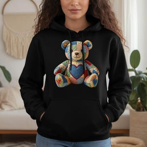 Pode incluir: Moletom preto com um design de urso de pelúcia patchwork colorido. O urso tem um coração azul e é feito de vários tecidos estampados. O moletom tem um bolso canguru e um capuz com cordão. Uma peça de roupa confortável e elegante.
