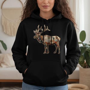 Könnte beinhalten: Schwarzer Kapuzenpullover mit einem Hirsch-Grafik. Der Hirsch ist mit einem Patchwork-Design in Braun-, Rot- und Cremetönen dargestellt. Der Kapuzenpullover hat eine Kapuze und eine Vordertasche. Der Hirsch hat ein großes Geweih.