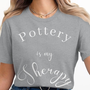 Könnte beinhalten: Graues T-Shirt mit dem Text "Pottery is my therapy" in weißen Buchstaben.