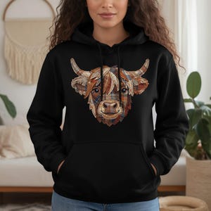 May include: Black hoodie featuring a detailed illustration of a Highland cow's head. The cow's face is a patchwork of brown, orange, and plaid patterns. The hoodie has a front pocket and a drawstring hood.