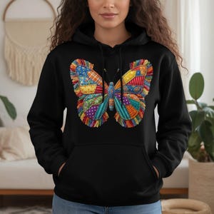 Könnte beinhalten: Schwarzer Kapuzenpullover mit einem bunten Schmetterlingsdesign. Der Schmetterling besteht aus Stoffflicken in Rot, Blau, Gelb und Grün. Der Hoodie hat eine Kapuze mit Kordelzug und eine Vordertasche.
