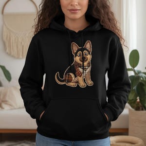 Puede incluir: Sudadera con capucha negra con un perro pastor alemán de dibujos animados con una bufanda a cuadros. El perro tiene colores marrón, negro y beige. La sudadera tiene un bolsillo delantero y una capucha con cordón. El gráfico del perro está centrado en la parte delantera.