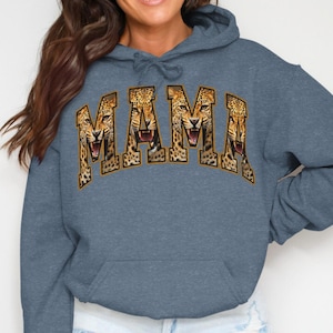 Puede incluir: Sudadera con capucha azul con la palabra "MAMA" en un diseño dorado y estampado de leopardo en el pecho. La sudadera tiene un bolsillo de canguro y una capucha con cordón. El diseño presenta una cara de leopardo.