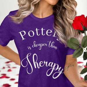 Könnte beinhalten: Lilafarbenes T-Shirt mit weißem Text, der "Töpfern ist günstiger als Therapie" lautet.