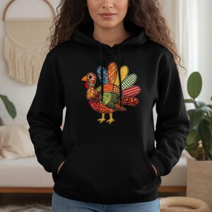 Puede incluir: Sudadera con capucha negra con un diseño de pavo patchwork colorido. El pavo tiene una carúncula roja, patas amarillas y plumas estampadas en azul, verde, rojo y amarillo. La sudadera tiene un bolsillo canguro.