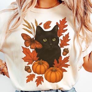 Pu&ograve; includere: T-shirt color avorio con un gatto nero circondato da elementi autunnali. Il design include zucche arancioni, foglie autunnali, una farfalla e ghiande. La maglietta &egrave; color crema, perfetta per l'autunno.