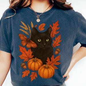 Pu&ograve; includere: T-shirt blu navy con un gatto nero circondato da foglie autunnali arancioni, zucche e una farfalla. Il design evoca un tema autunnale, perfetto per la stagione.