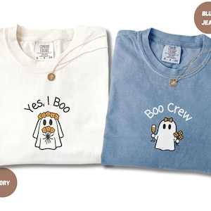 May include: Two t-shirts, one ivory and one blue jean, with ghost graphics. The ivory shirt says "Yes, I Boo" and the blue jean shirt says "Boo Crew". Both shirts have a Comfort Colors tag and a gold necklace.