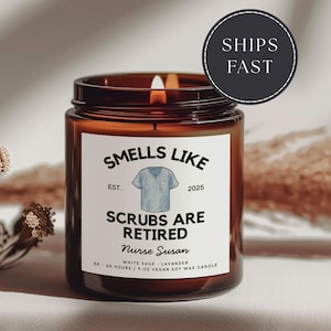 May include: A brown glass candle jar with a lit wick. The label reads "Smells Like Scrubs Are Retired Nurse Susan" with a blue scrubs graphic. The candle is 9 oz and made of vegan soy wax.
