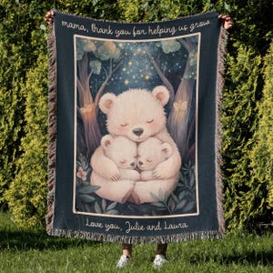 The image shows a person holding up a woven tapestry or blanket. 

The tapestry features a central illustration of three stylized polar bears; a larger bear embracing two smaller ones. The bears are depicted in a serene, nighttime forest setting under a starry sky. The overall aesthetic is gentle, warm, and slightly whimsical. 
At the top of the tapestry, a message reads, "mama, thank you for helping us grow." At the bottom, a personalized inscription says, "Love you, Julie and Laura."