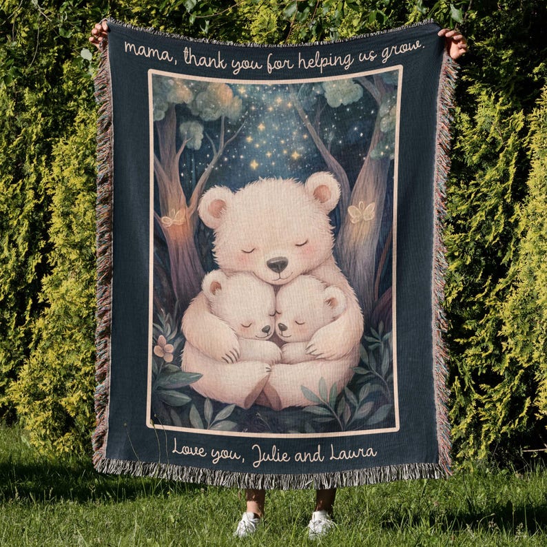 The image shows a person holding up a woven tapestry or blanket. 

The tapestry features a central illustration of three stylized polar bears; a larger bear embracing two smaller ones. The bears are depicted in a serene, nighttime forest setting under a starry sky. The overall aesthetic is gentle, warm, and slightly whimsical. 
At the top of the tapestry, a message reads, "mama, thank you for helping us grow." At the bottom, a personalized inscription says, "Love you, Julie and Laura."