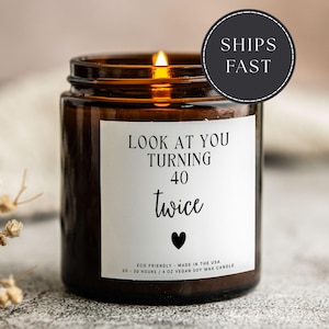 May include: A lit brown glass candle jar with a white label. The label reads "LOOK AT YOU TURNING 40 twice" with a black heart. The candle is made with vegan soy wax and has a burn time of 20-30 hours. The text "SHIPS FAST" is in a black circle.