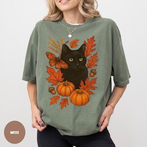 Pu&ograve; includere: Una maglietta verde muschio con un disegno di un gatto nero circondato da foglie autunnali arancioni, zucche e ghiande. Sono inclusi anche una farfalla marrone e spighe di grano. La maglietta ha una vestibilit&agrave; ampia.