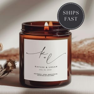 May include: A lit amber glass candle with a white label featuring the names "Kaylee & Logan" and the date "July 18, 2026". The candle is 4oz and made with natural soy wax. The text "SHIPS FAST" is in a black circle.
