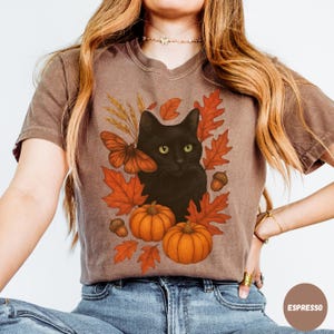 Pu&ograve; includere: T-shirt color espresso con un design di gatto nero circondato da elementi autunnali. La grafica include zucche arancioni, foglie autunnali, una farfalla e ghiande. La maglietta ha uno scollo a V e maniche corte.