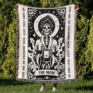 May include: Black and white woven tapestry with a skeleton in a robe holding a coffee mug. The tapestry has the text "YOU ARE AN AWESOME MOM. KEEP THAT SHIT UP." and "THE MOM" on it.