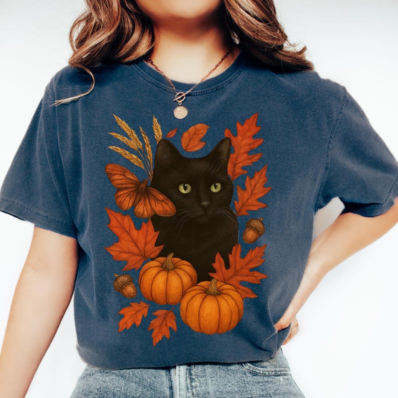Pu&ograve; includere: T-shirt blu navy con un gatto nero circondato da foglie autunnali arancioni, zucche e una farfalla. Il design evoca un tema autunnale, perfetto per la stagione.