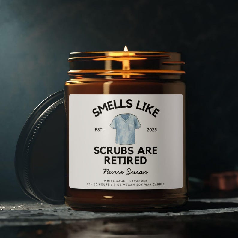 May include: A brown glass jar candle with a lit wick and a black lid. The label reads "Smells Like Scrubs Are Retired Nurse Susan" with a blue scrub top graphic. The candle is a 9 oz vegan soy wax candle.