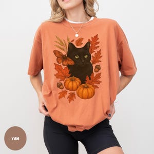 Pu&ograve; includere: T-shirt arancione con un disegno di gatto nero circondato da foglie autunnali, zucche e una farfalla. La grafica ha un tema autunnale, con colori caldi e illustrazioni dettagliate. La maglietta &egrave; uno stile casual e confortevole.