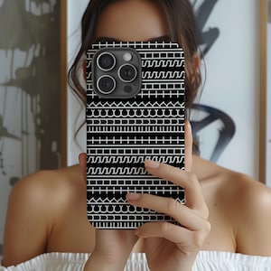 May include: Black phone case with a repeating white geometric pattern. The case has a camera cutout and is held in front of a person. The design includes lines, curves, and small rectangles, creating a textured appearance.