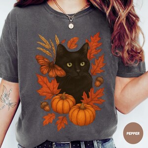 Pu&ograve; includere: T-shirt grigio scuro con un gatto nero circondato da foglie autunnali arancioni, zucche e una falena. Il design include spighe di grano e ghiande, evocando un tema autunnale. La parola "PEPPER" &egrave; stampata in basso a destra.