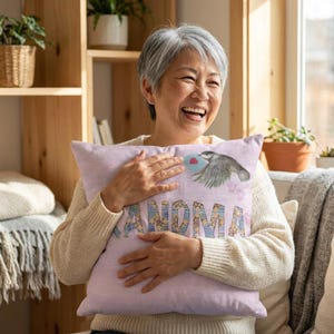 Puede incluir: Un cojín rosa claro con la palabra "GRANDMA" en letras de patchwork. El cojín presenta una ilustración de acuarela de un pájaro que lleva una carta en forma de corazón. El cojín está siendo sostenido por una persona.