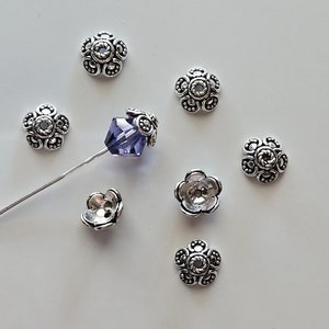 May include: Silver flower-shaped bead caps with a textured design. The caps are arranged on a white surface.