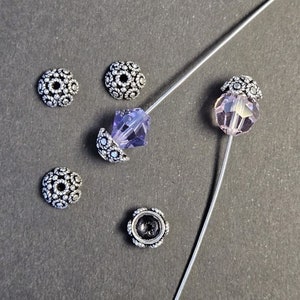 May include: Close-up of jewelry-making components. The image features several silver-colored, flower-shaped bead caps and two faceted beads, one purple and one pink, strung on a thin wire. The beads and caps are arranged on a dark gray surface.