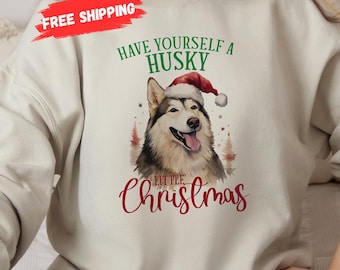 Puede incluir: Sudadera color crema con una ilustración en acuarela de un perro Husky con un gorro de Papá Noel. El texto "Have Yourself a Husky Little Christmas" está impreso encima y debajo de la imagen del perro en verde y rojo.