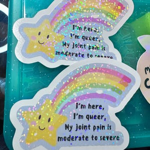 Puede incluir: Dos pegatinas con un diseño de estrella fugaz. La estrella amarilla tiene una cara sonriente, y la estela del arcoíris es brillante. El texto en las pegatinas dice: "I'm here, I'm queer, My joint pain is moderate to severe."