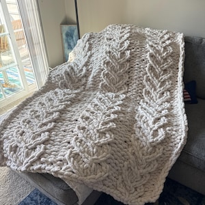 May include: A chunky knit blanket in a light gray color, draped over a gray sofa. The blanket features a cable knit design, creating a textured and cozy appearance. The soft, plush material suggests warmth and comfort, perfect for home decor.