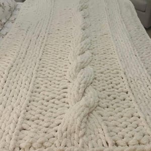 May include: A large, off-white chunky knit blanket with a cable knit design. The blanket is made of thick, soft yarn and is spread out on a bed. The cable knit pattern runs down the center of the blanket, with a textured knit on either side.