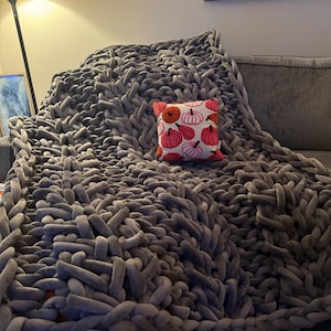 May include: A large, chunky knit gray blanket draped over a gray sofa. A small square pillow with a pumpkin pattern in orange, pink, and white rests on the blanket. The blanket appears soft and cozy, perfect for snuggling.