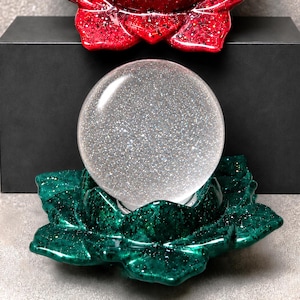 May include: A clear, spherical glass orb filled with fine silver glitter, resting in a dark green, multi-petaled lotus flower base. A second, identical lotus flower base in a deep red color is positioned directly above the orb.