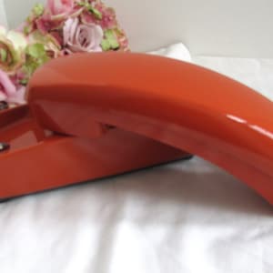May include: A vintage orange rotary phone with a coiled cord. The phone has a sleek, modern design and is in excellent condition.
