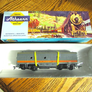 May include: Athearn model train in a box. The box features a colourful illustration of a steam train and the text "TRAINS in MINIATURE". The model train is grey with orange and yellow accents, secured in a clear plastic case.