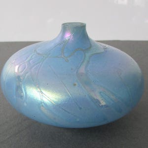 May include: A small, round, iridescent blue glass vase. The vase has a short neck and a bulbous body. The surface has a subtle, swirling pattern. The vase is approximately 4 inches in diameter.