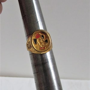 以下が含まれることがあります： 赤い帽子と赤い蝶ネクタイをした漫画の犬が描かれた金色の指輪。指輪には「Muttley's Adventures」という文字が刻まれています。