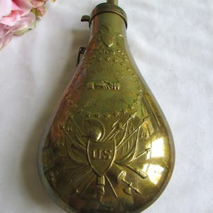 May include: Vintage brass powder flask, teardrop-shaped, with engraved patriotic motifs. It features an eagle, stars, and "US" on a shield. The flask has a spout and hinged top, likely for storing and dispensing gunpowder. A historical item.
