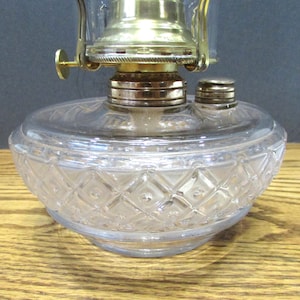 Puede incluir: Una lámpara de aceite de vidrio transparente con un patrón de diamante en la base. La lámpara tiene un quemador de latón y una chimenea de vidrio transparente.
