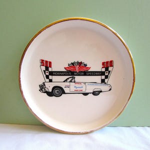 May include: A round white ceramic plate with a gold-toned rim featuring a graphic of a white Plymouth pace car. The design includes the text Indianapolis Motor Speedway, the number 500, and a winged emblem centered above the car illustration.