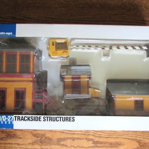 Puede incluir: Una caja de estructuras de vía férrea modelo HO. El conjunto incluye una torre amarilla, un edificio amarillo con techo rojo, un edificio amarillo con techo marrón y una barrera de cruce amarilla.