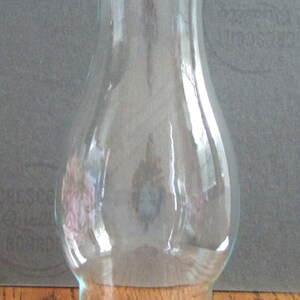 May include: A clear glass lamp chimney with a wide base and a narrow top. The chimney is designed to fit over a lamp wick and is used to protect the flame from drafts.
