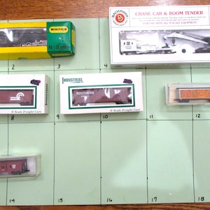 Könnte beinhalten: Eine Sammlung von Modellbahnwagen und Zubehör, darunter ein Kranwagen, Güterwagen und eine Lokomotive. Die Artikel sind in Kartons und Vitrinen verpackt, mit verschiedenen Farben und Designs. Der Maßstab ist N.